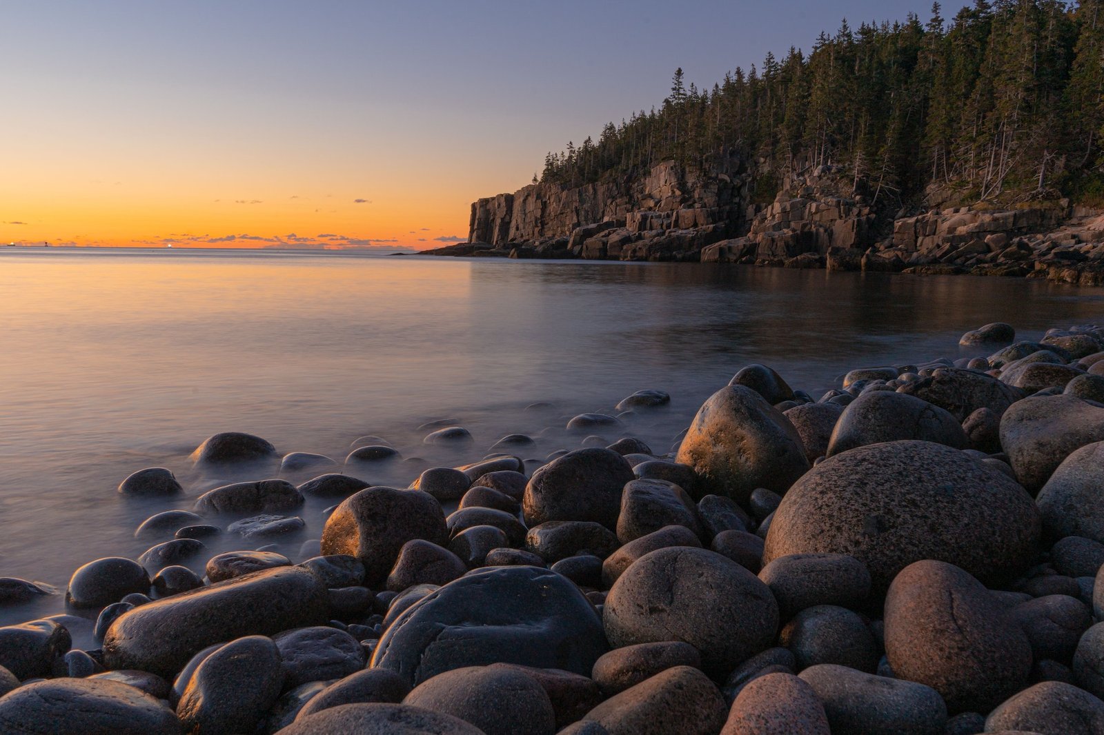 Beautiful scene of Boulder Beach in Acadia National Park, Maine with green hills and an orange sky